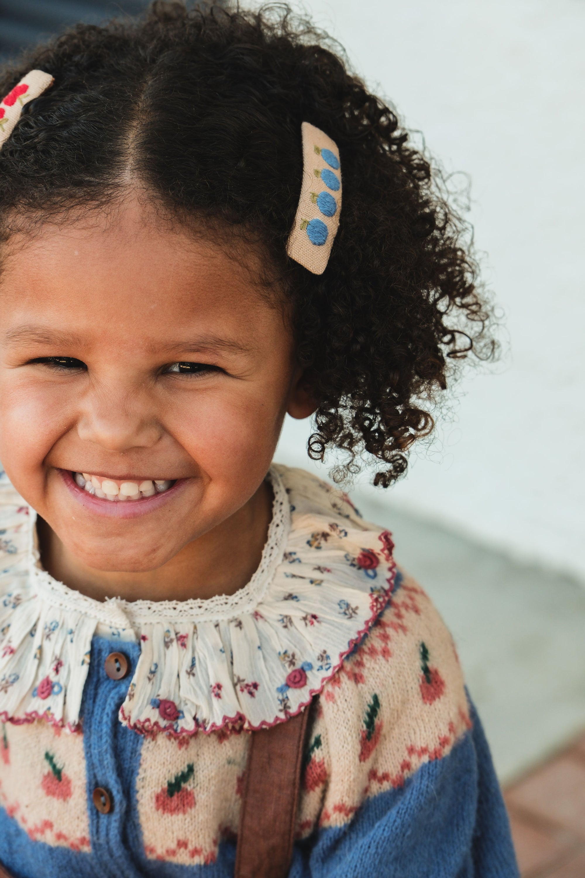 Child with curly hair wearing a floral dress and blue cardigan, smiling outdoors.