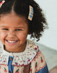 Child with curly hair wearing a floral dress and blue cardigan, smiling outdoors.